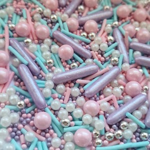 May include: A close-up of a mix of colourful sprinkles. The sprinkles include light pink spheres, iridescent purple rods, silver dragees, light blue sticks, and small white and purple spheres. The sprinkles are scattered and create a textured surface.