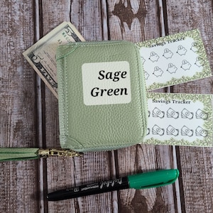Op de afbeelding: Sagegroene rits portemonnee met een gouden rits trekkoord en een groene kwast. De portemonnee heeft een gepersonaliseerd naamplaatje met de tekst "Sage Green". De portemonnee wordt geleverd met twee vellen stickers met de tekst "Savings Tracker" en afbeeldingen van spaarpotten en stapels geld.