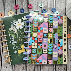 May include: Two decorative planners with floral designs. One planner has a dark green cover with white flowers and the words "Find Beauty". The other planner has a colorful geometric floral pattern. Gold and blue disc-bound rings.