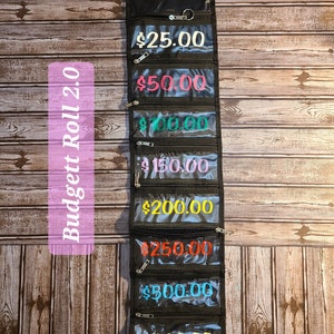 May include: A black fabric roll-up budget organiser with clear plastic windows displaying pound sterling amounts: £25.00, £50.00, £100.00, £150.00, £200.00, £250.00, £500.00, and £1000.00.