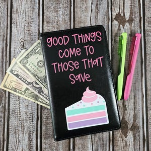 May include: Black leather checkbook cover with the text "Good Things Come To Those That Save" in pink lettering. A pink and green layered cake with white frosting is on the cover.