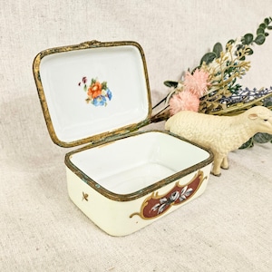 May include: A white porcelain trinket box with a gold trim and floral design on the lid. The box is open and the inside is white with a red and gold design.