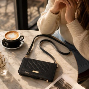 May include: Black crossbody wallet with a quilted design and a gold-colored accent. The wallet has a long strap and a zipper closure. A cup of coffee with latte art sits on a table next to the wallet.
