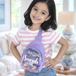 Puede incluir: Un bolso bandolera de color lavanda con un bolsillo frontal transparente lleno de lentejuelas de colores en forma de estrellas y flores. El nombre Sarah está impreso en letras blancas sobre el bolsillo transparente, con una correa a juego.