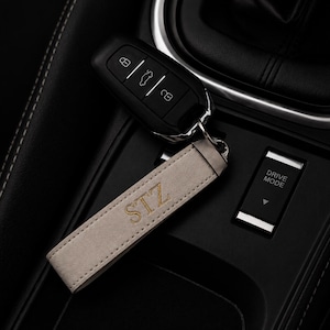 May include: A car key and a light grey leather keyring with the embroidered initials "STZ" in gold. The keyring is attached to the car key. The car key is black. The background is a car's interior.