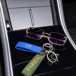 Puede incluir: Gafas de sol rectangulares sin montura con lentes tintadas en morado y marcos dorados, situadas sobre dos llaveros de cuero. Uno es azul con las letras ZR en dorado y el otro es verde oliva con el nombre FARIDA grabado en dorado.