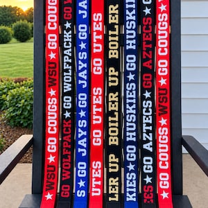 May include: Eight vertical fabric straps with gold-tone metal swivel clasps at the bottom. The straps feature various team slogans and names in white or gold text, including GO COUGS, GO WOLFPACK, GO JAYS, GO UTES, BOILER UP, and GO HUSKIES.