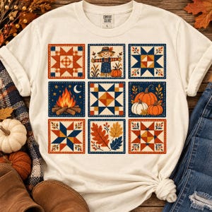 May include: A cream-colored t-shirt featuring a grid of nine square illustrations. The designs include a scarecrow, a campfire, pumpkins, autumn leaves, and various geometric quilt-style star patterns in shades of orange, blue, and yellow.
