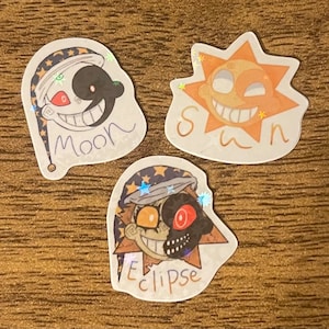 Puede incluir: Tres pegatinas holográficas con caras de dibujos animados de la luna, el sol y el eclipse. La luna es blanca con un ojo negro y una boca roja. El sol es naranja con un ojo amarillo y una boca roja. El eclipse es blanco y negro con un ojo amarillo y una boca roja. Cada pegatina tiene el nombre del cuerpo celeste escrito debajo de la imagen.