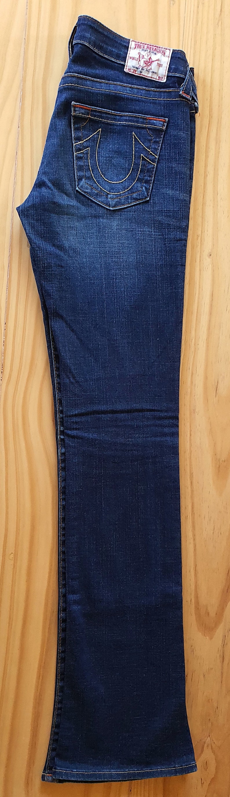 May include: A pair of dark blue denim jeans with a bootcut leg. The jeans have a red and white True Religion brand label on the back pocket.