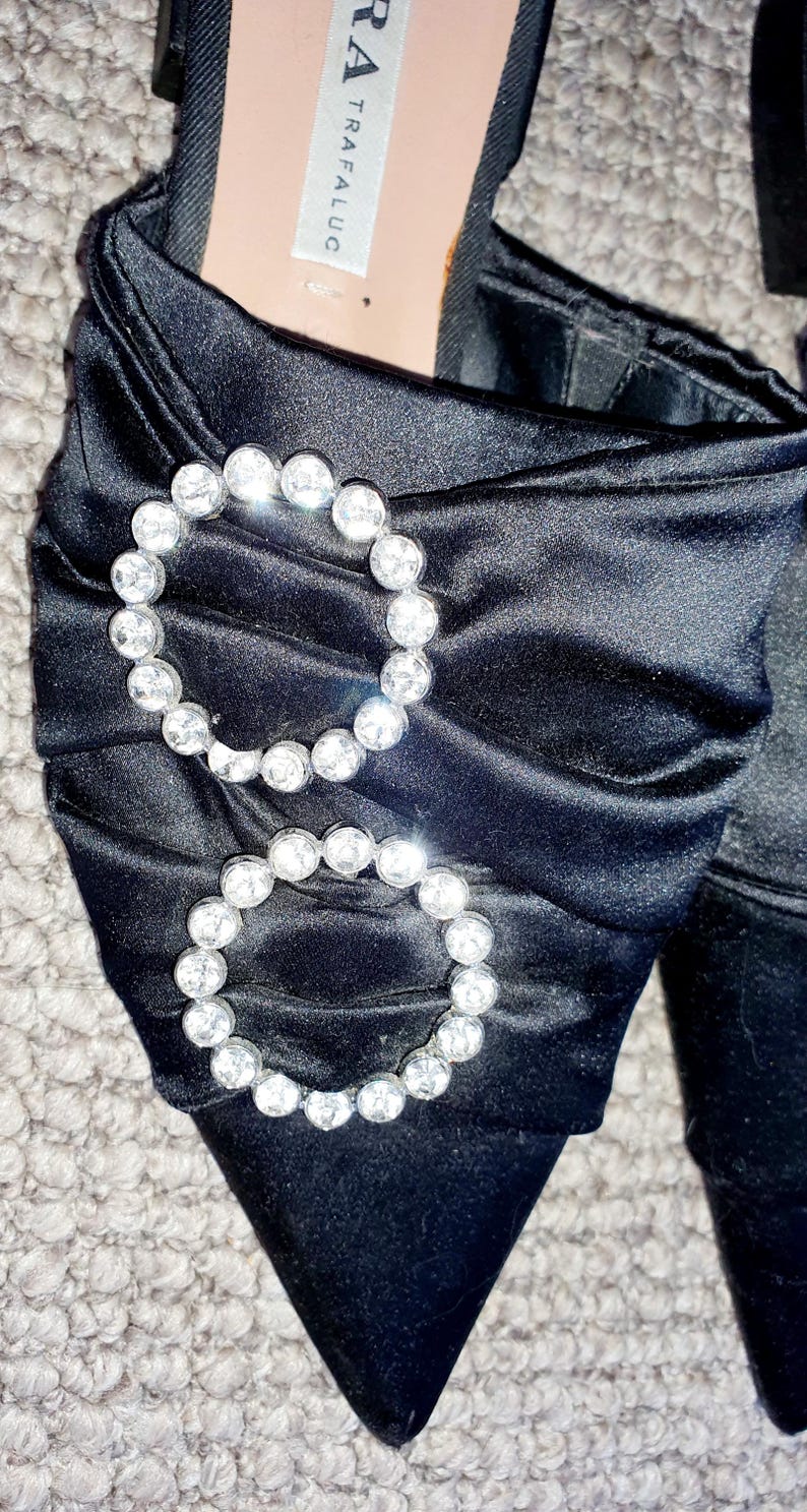 Puede incluir: Zapatos de tac&oacute;n de sat&eacute;n negro con punta puntiaguda y dos grandes adornos de pedrer&iacute;a. Los zapatos tienen una etiqueta que dice "TRAFA LUC" y "ZARA".