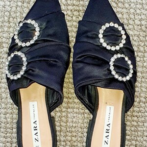 Puede incluir: Un par de mules de tac&oacute;n de sat&eacute;n negro con punta puntiaguda. Los zapatos presentan dos grandes hebillas redondas adornadas con pedrer&iacute;a. Los zapatos est&aacute;n etiquetados como "ZARA TRAFALUC 38".