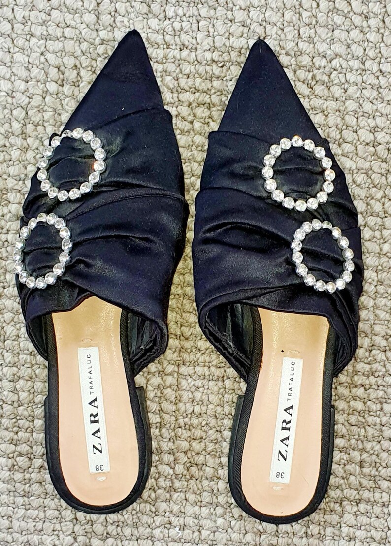 Puede incluir: Un par de mules de tac&oacute;n de sat&eacute;n negro con punta puntiaguda. Los zapatos presentan dos grandes hebillas redondas adornadas con pedrer&iacute;a. Los zapatos est&aacute;n etiquetados como "ZARA TRAFALUC 38".