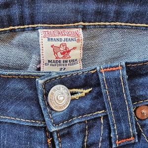 May include: Close-up of a pair of blue denim jeans with a True Religion brand label and a metal button. The label reads "True Religion Brand Jeans" and "Made in USA of Imported Fabric 27".