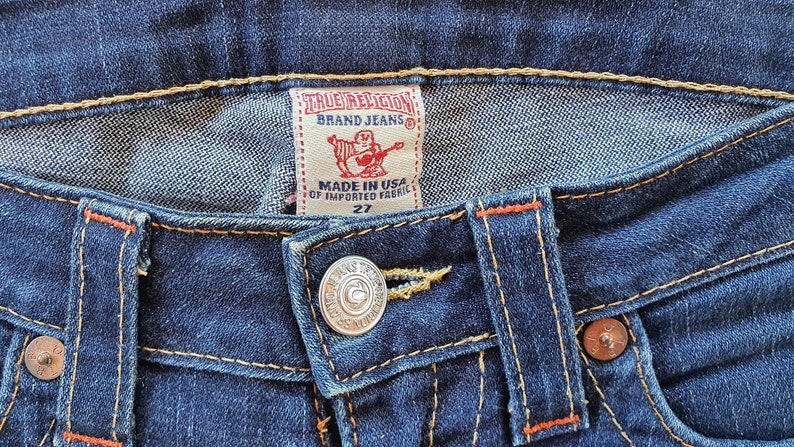 May include: Close-up of a pair of blue denim jeans with a True Religion brand label and a metal button. The label reads "True Religion Brand Jeans" and "Made in USA of Imported Fabric 27".