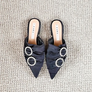 Puede incluir: Un par de mules de sat&eacute;n negro de punta con un detalle de hebilla adornada con joyas. Los zapatos est&aacute;n etiquetados con "ZARA" en la plantilla.