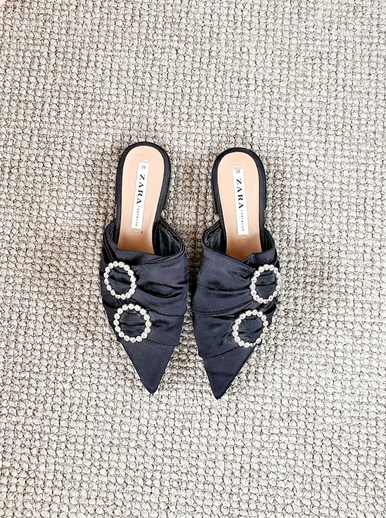 Puede incluir: Un par de mules de sat&eacute;n negro de punta con un detalle de hebilla adornada con joyas. Los zapatos est&aacute;n etiquetados con "ZARA" en la plantilla.