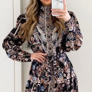Puede incluir: Un vestido camisa de manga larga con estampado negro y marrón, cuello alto, cierre de botones y cinturón. El vestido tiene un diseño floral y geométrico.