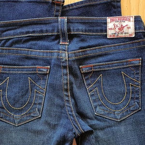 May include: A pair of dark blue denim jeans with a red stitching detail. The back pockets have a distinctive yellow embroidered horseshoe logo with the text "True Religion" on the right back pocket.