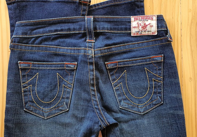 May include: A pair of dark blue denim jeans with a red stitching detail. The back pockets have a distinctive yellow embroidered horseshoe logo with the text "True Religion" on the right back pocket.