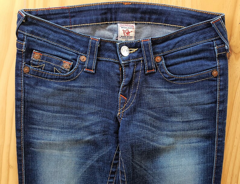 May include: A pair of blue denim jeans with orange stitching and a True Religion brand label. The jeans have a faded look and a classic five-pocket style.