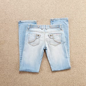 Peut inclure: Jean clair en denim avec une conception classique à cinq poches. Le jean a une taille basse et un léger évasement en bas. Les poches arrière ont un design incurvé unique. Le jean est posé à plat sur une surface de couleur neutre.