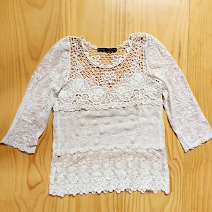 Peut inclure: Un haut en dentelle blanche avec des manches trois-quarts. Le haut présente une encolure en crochet et un design superposé avec des motifs floraux et géométriques complexes. Le tissu délicat crée un effet semi-transparent.