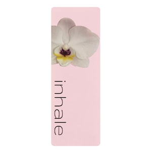 May include: A rectangular light pink yoga mat featuring a white orchid flower with a yellow and dark purple centre at the top. The word inhale is printed in a simple black sans-serif font vertically along the left side of the mat surface.
