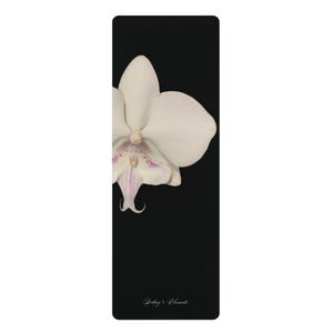 以下が含まれることがあります： 無地の黒い背景に、左側に白い蘭の花の一部が描かれた長方形のヨガマット。花の中心付近には淡い紫色の模様があります。下部には白い文字でDestiny's Elementsと印刷されています。