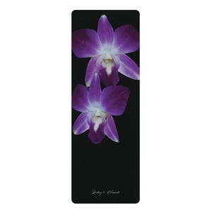 Puede incluir: Una esterilla de yoga rectangular negra con dos orquídeas púrpuras vibrantes con centros blancos situadas en la parte superior. El texto Destiny's Elements está impreso en una fuente cursiva blanca pequeña y elegante en la parte inferior central.