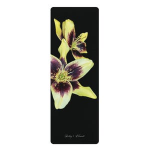 Puede incluir: Una esterilla de yoga rectangular negra con un estampado botánico de dos lirios con pétalos de color amarillo pálido y centros de color púrpura oscuro. El texto "Destiny's Elements" está impreso en una pequeña y elegante fuente blanca.