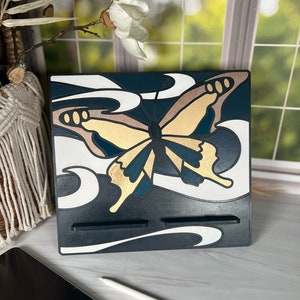 Può includere: Un supporto per telefono in legno nero con un design a farfalla bianco e oro. Il supporto ha due slot per tenere i telefoni o altri dispositivi.