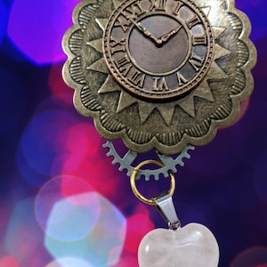 May include: A steampunk-style pendant featuring a bronze-colored clock face with Roman numerals, set within a decorative, scalloped frame. A small gear and a gold-colored ring connect the clock to a heart-shaped, pale pink gemstone pendant.