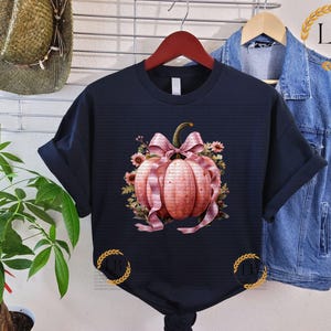 May include: A black t-shirt with a pink and white pumpkin with a bow and floral accents. The pumpkin is surrounded by leaves and vines.
