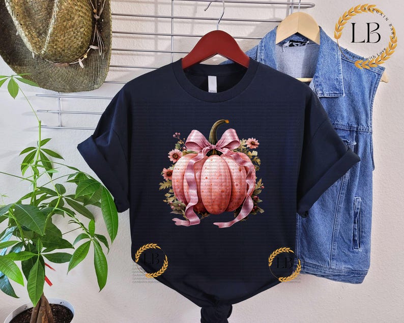 May include: A black t-shirt with a pink and white pumpkin with a bow and floral accents. The pumpkin is surrounded by leaves and vines.