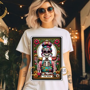 May include: A tarot card illustration featuring a skeleton wearing a colorful floral headdress and a blue and green apron. The card is titled "The Mom" and is surrounded by flowers and other decorative elements.