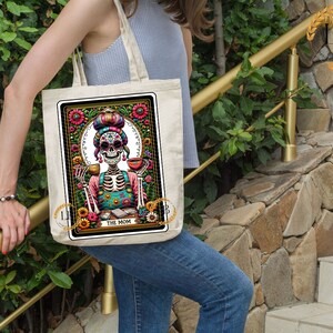 May include: A white tote bag with a colorful illustration of a skeleton wearing a floral headband and holding a cup. The illustration is surrounded by a floral border and the text "The Mom" is printed below it.