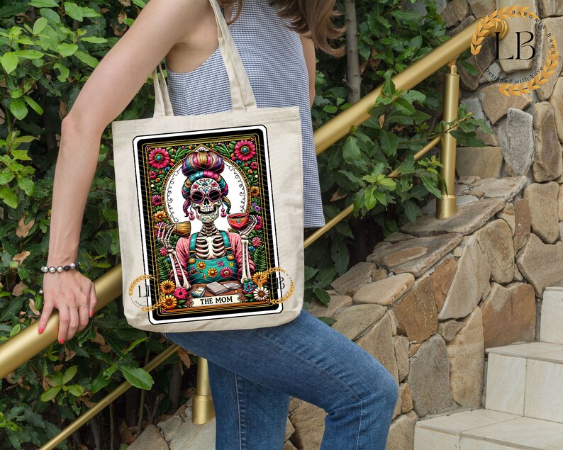 May include: A white tote bag with a colorful illustration of a skeleton wearing a floral headband and holding a cup. The illustration is surrounded by a floral border and the text "The Mom" is printed below it.