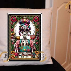 May include: A white throw pillow with a colorful illustration of a skeleton woman with flowers and a cup of tea. The illustration is framed in a black border with gold accents. The text "THE MOM" is at the bottom of the illustration.