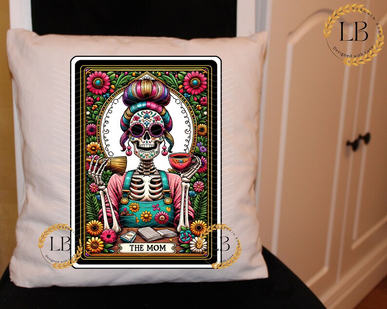 May include: A white throw pillow with a colorful illustration of a skeleton woman with flowers and a cup of tea. The illustration is framed in a black border with gold accents. The text "THE MOM" is at the bottom of the illustration.