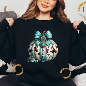 May include: A black sweatshirt with a teal and gold pumpkin design. The pumpkin has a teal bow with a gold buckle and is decorated with gold and white details.