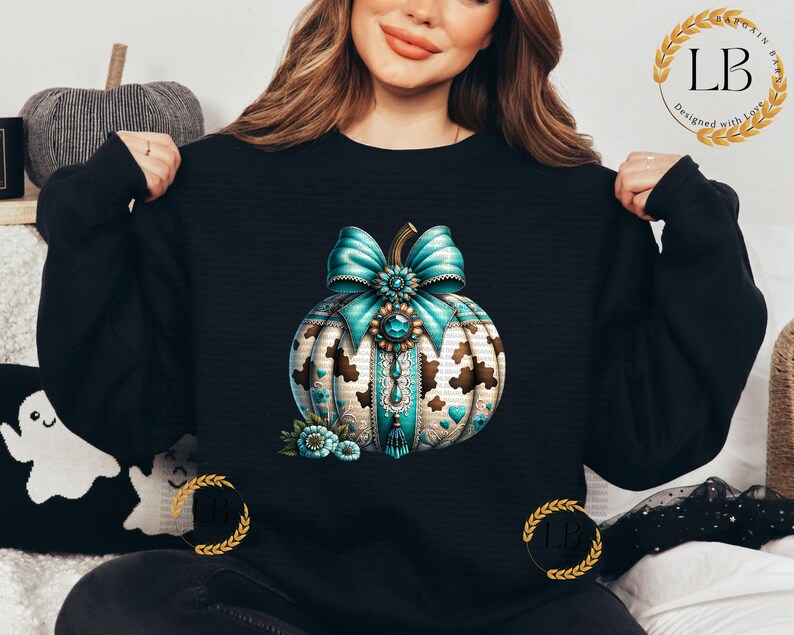 May include: A black sweatshirt with a teal and gold pumpkin design. The pumpkin has a teal bow with a gold buckle and is decorated with gold and white details.