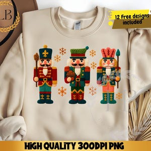 Könnte beinhalten: Beiges Sweatshirt mit einem gestickten Design von drei bunten Nussknackern. Jeder Nussknacker trägt eine andere Uniform mit goldenen Akzenten und hält einen Stab oder einen Leckerbissen. Der Text "12 Free designs included" befindet sich in der oberen rechten Ecke.