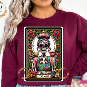 May include: A maroon sweatshirt with a colorful graphic print of a skeleton woman with a flower crown and a cup of coffee. The graphic is in the style of a tarot card and says "The Mom" at the bottom.