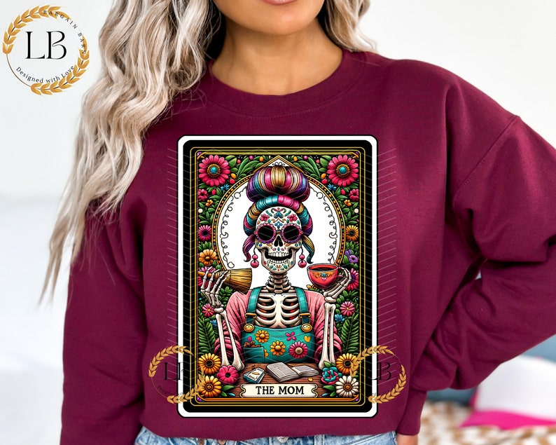 May include: A maroon sweatshirt with a colorful graphic print of a skeleton woman with a flower crown and a cup of coffee. The graphic is in the style of a tarot card and says "The Mom" at the bottom.