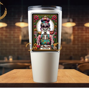 May include: White stainless steel tumbler with a colorful illustration of a skeleton wearing a floral dress and holding a cup. The illustration is framed with a gold border and the text "The Mom" is printed below the image.