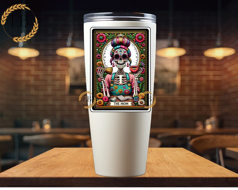 May include: White stainless steel tumbler with a colorful illustration of a skeleton wearing a floral dress and holding a cup. The illustration is framed with a gold border and the text "The Mom" is printed below the image.