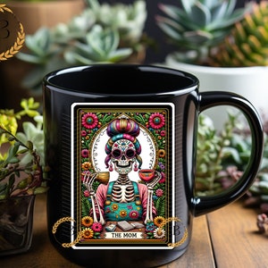 May include: Black coffee mug with a colorful illustration of a skeleton woman with a floral background. The illustration is in the style of a tarot card and says "The Mom" at the bottom.