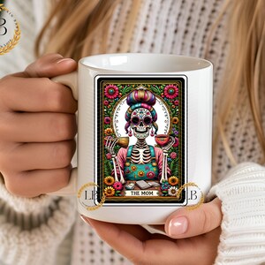 May include: White ceramic mug with a colorful illustration of a skeleton woman wearing a floral headband and holding a cup. The illustration is framed with a black border and floral designs. The text "THE MOM" is printed below the illustration.