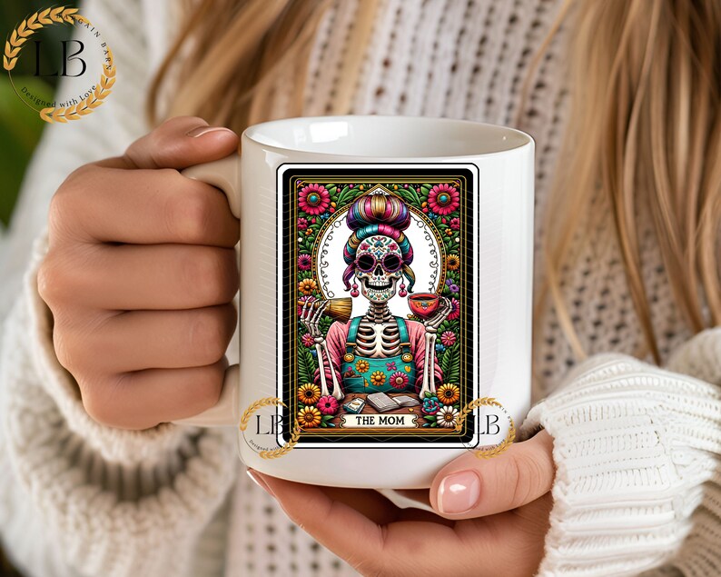 May include: White ceramic mug with a colorful illustration of a skeleton woman wearing a floral headband and holding a cup. The illustration is framed with a black border and floral designs. The text "THE MOM" is printed below the illustration.